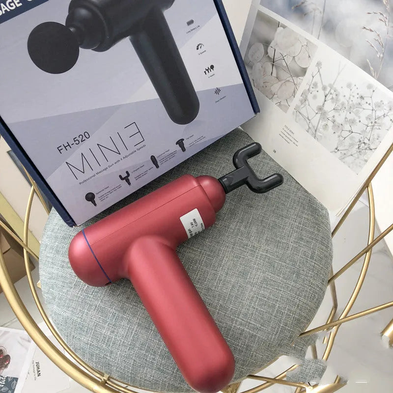 Grey, red, and black handheld fascia gun massager on a table with USB charging cable.