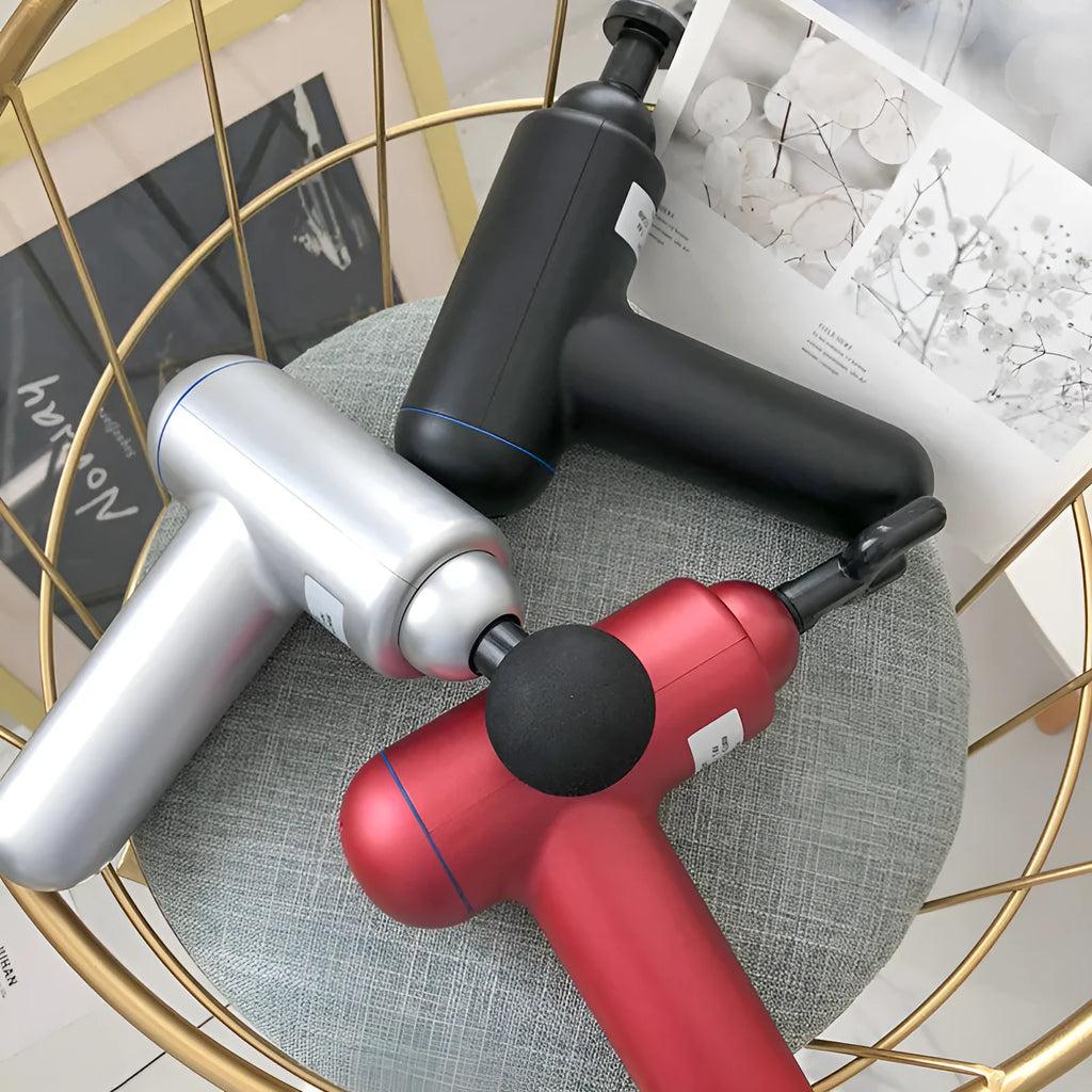 Grey, red, and black handheld fascia gun massager on a table with USB charging cable.