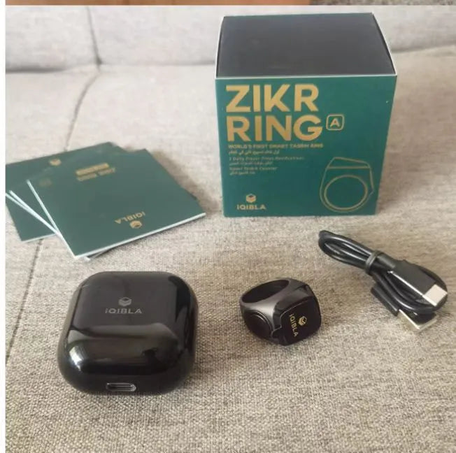 Silver smart ring with a small OLED display showing prayer count, next to its USB charging case.