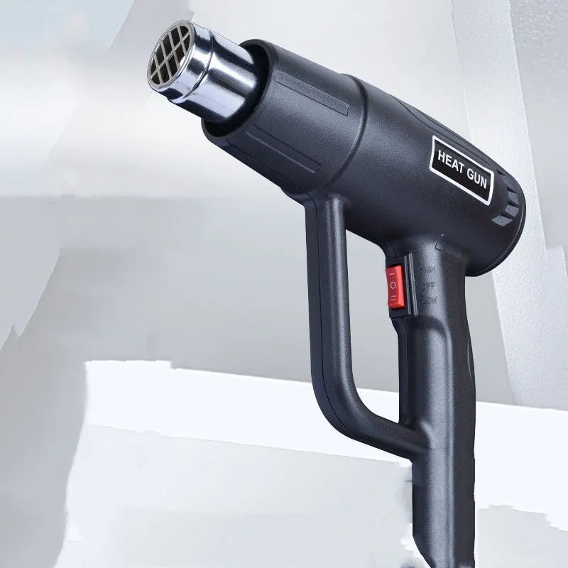 Black industrial hot air gun with a two-speed switch and grid-pattern nozzle on a workbench.