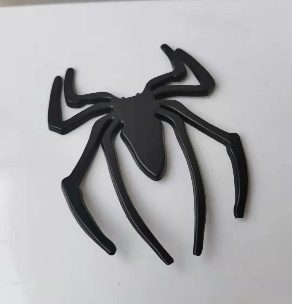 Three-dimensional zinc alloy spider bumper sticker in metallic colours on a surface.